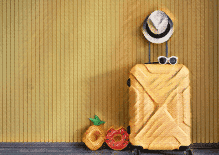 yellow suitcase with hat on handle and inflatable pineapple and donut sitting beside suitcase in front of a yellow wall